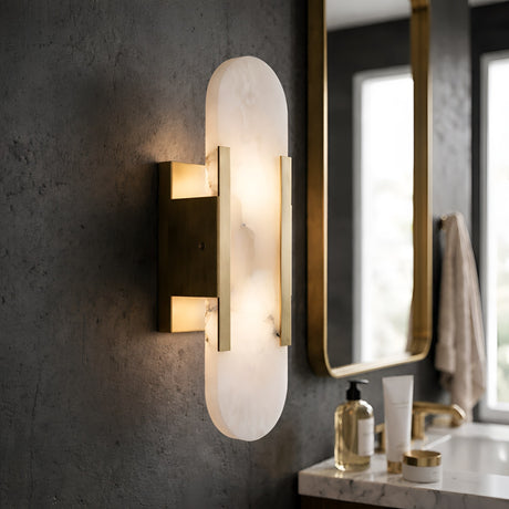 The Lirone 1 Light Small Vertical Capsule Alabaster Wall Light in soft white and brass is mounted on a textured dark wall near a bathroom vanity, paired with a gold-framed mirror, hand soap, and a towel in the background.