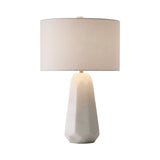 The Averon 1 Light Faceted Alabaster Table Lamp features a sculptural brass and soft white base with a large, round off-white fabric shade, shown here against a plain white background.