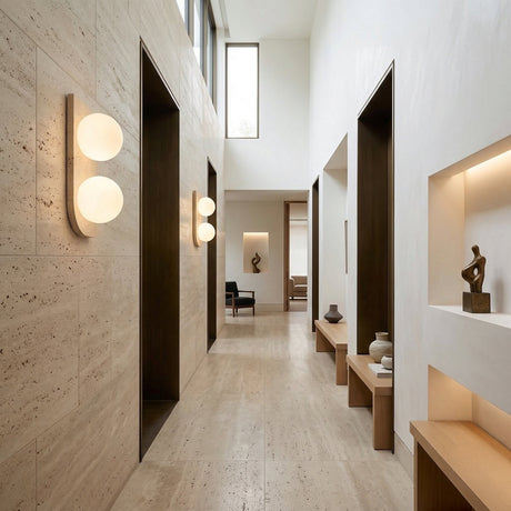 A modern hallway with beige stone floors and walls features Virella 2 Light Vertical Travertine Wall Lights in light beige for ambient lighting, minimalist benches, art sculptures on shelves, and tall windows that let in natural light.