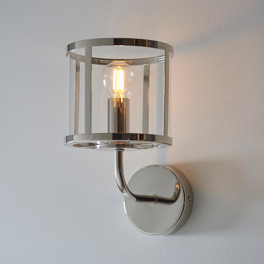 Ellery Wall Light in nickel with cylindrical clear glass shade, metal frame, and curved arm on a round backplate, shown lit against a white wall.