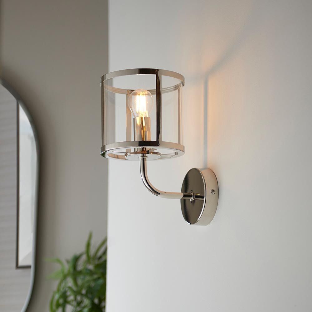 Ellery Wall Light in nickel with a cylindrical clear glass shade and exposed bulb, mounted on a light wall in a hallway setting.