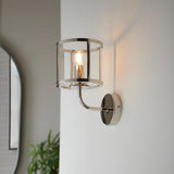 Ellery Wall Light in nickel with a cylindrical clear glass shade and exposed bulb, mounted on a light wall in a hallway setting.