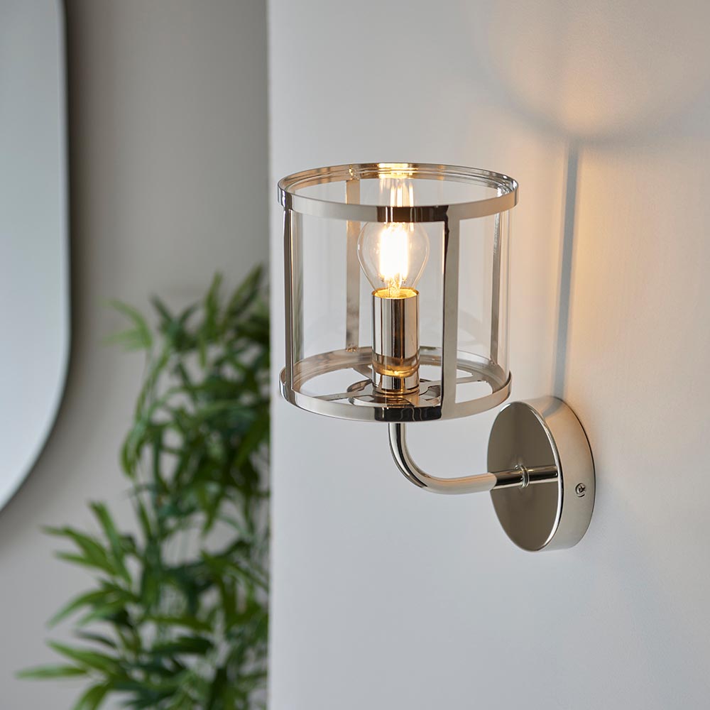 Ellery Wall Light in nickel with a cylindrical clear glass shade and exposed bulb, mounted on a plain wall beside a green plant.