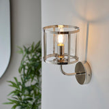 Ellery Wall Light in nickel with a cylindrical clear glass shade and exposed bulb, mounted on a plain wall beside a green plant.