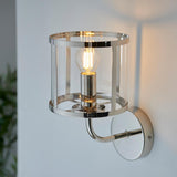 Ellery Wall Light in nickel with a cylindrical clear glass shade and exposed bulb, mounted on a white wall via a curved arm and round backplate.