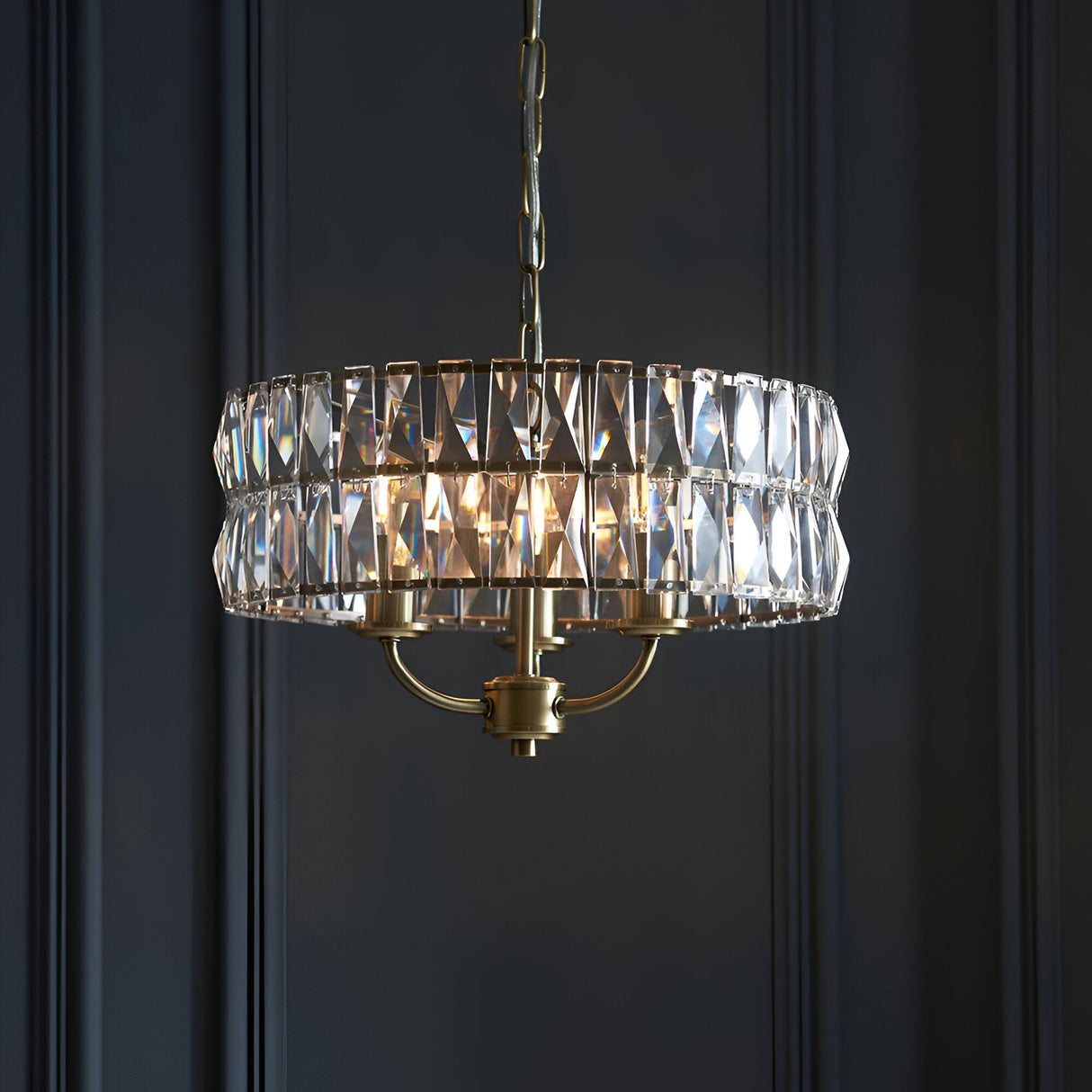 Endon Bevmont 3 light pendant in brass with faceted crystal glass drum shade on a chain suspension, shown against a dark panelled wall.