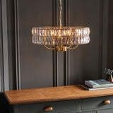Bevmont 5 Light Pendant in brass with a drum-shaped frame of faceted crystal glass, hung on a chain above a wooden sideboard in a dark panelled room.