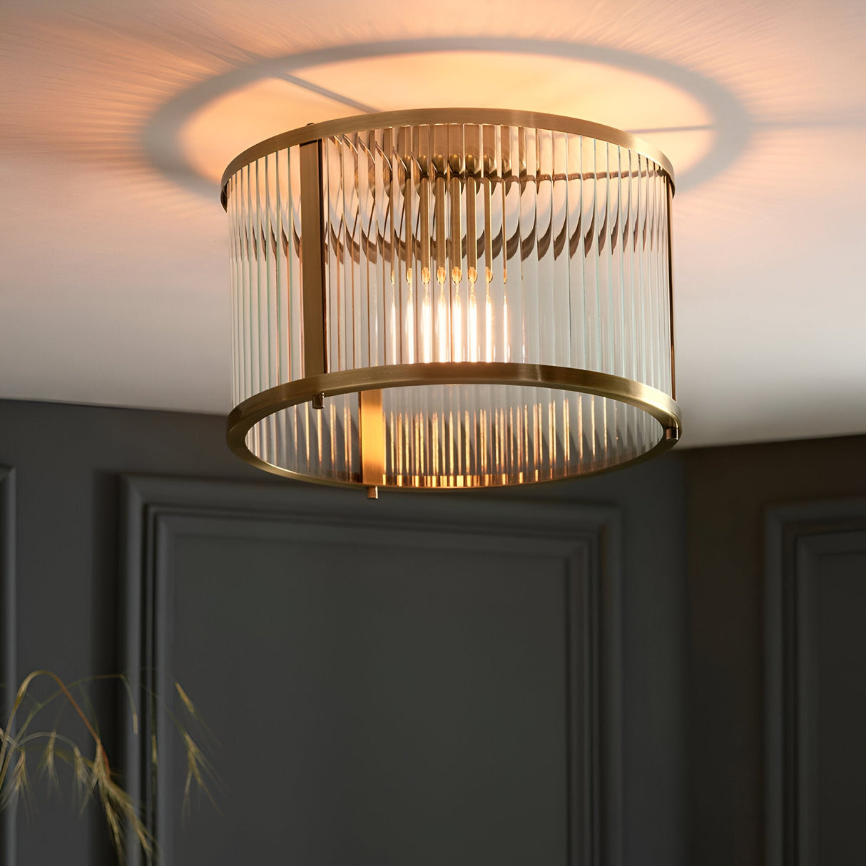 Rothbury flush ceiling light in antique brass, round drum shade with vertical glass rods and single lit bulb, mounted on a dark panelled wall ceiling.