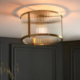 Rothbury flush ceiling light in antique brass, round drum shade with vertical glass rods and single lit bulb, mounted on a dark panelled wall ceiling.