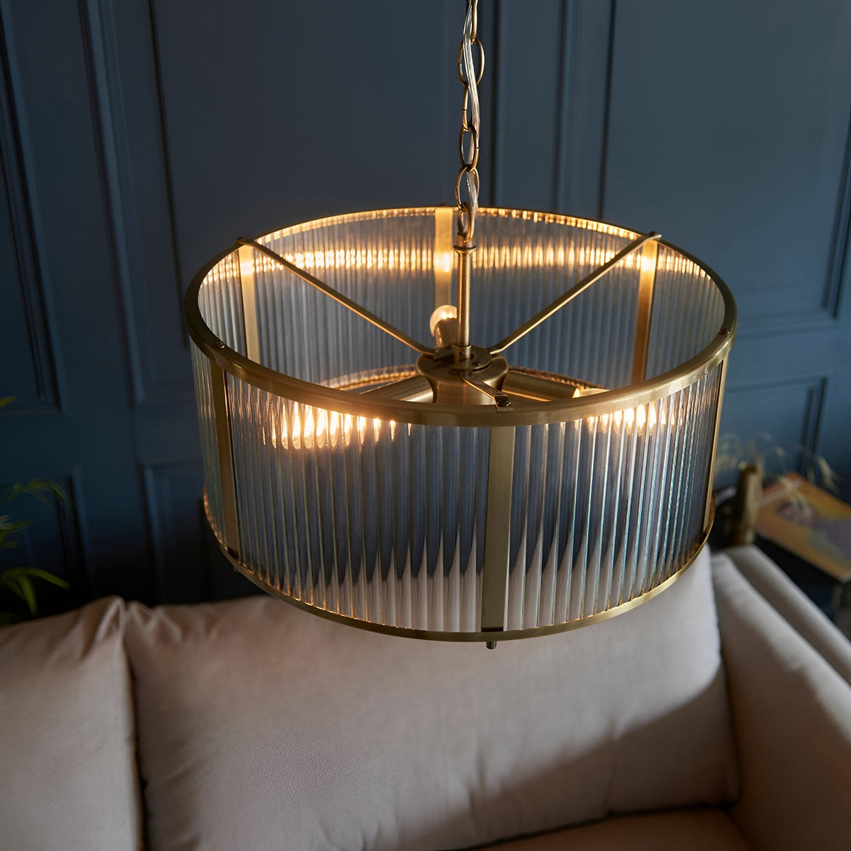 Rothbury 3 Light Pendant in antique brass with ribbed glass panel shades, hung on a chain above a beige sofa in a living room setting.