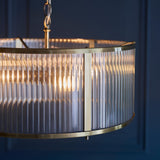 Close-up of the Rothbury 3 Light Pendant in antique brass, showing ribbed glass shade panels and brass frame detail against a dark blue wall.
