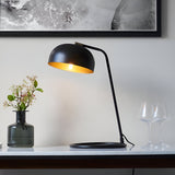 Ashdon Table Lamp in matt black metal with an antique brass inner shade, lit on a marble sideboard beside a vase and glassware.
