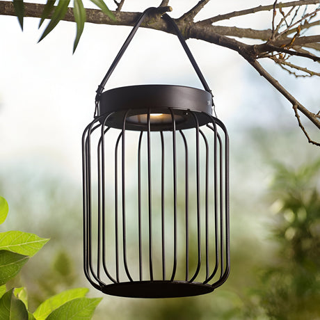 Endon Cranwood LED solar table lamp in matt black, open metal cage design with leather strap handle, hung from a tree branch in a garden setting.