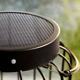 Close-up of the Endon Cranwood LED solar table lamp showing its matt black metal cage top and solar panel, set outdoors against soft greenery.