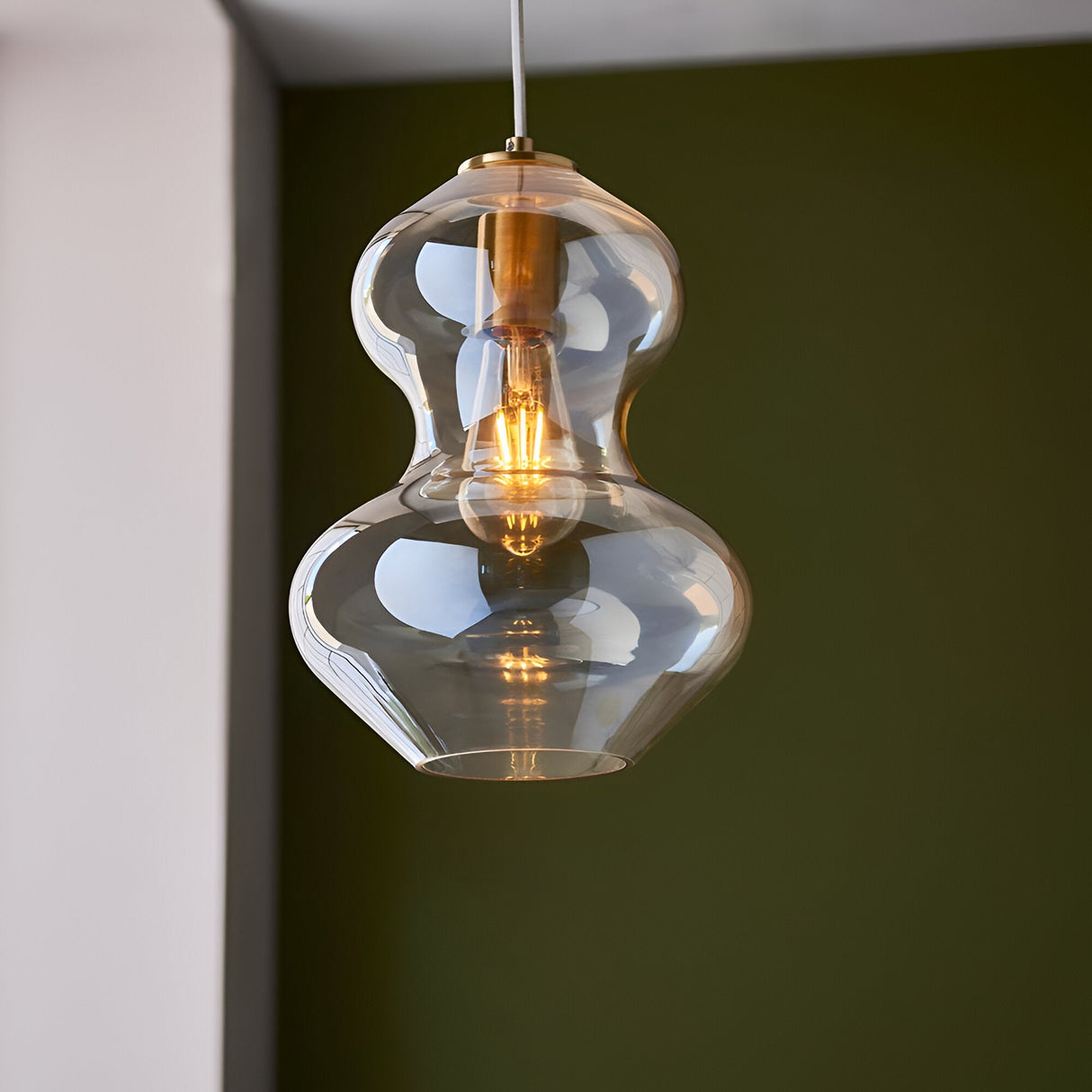 Brockley Pendant in champagne lustre glass with a curved double-bubble shade and satin brass fitting, shown lit against a dark green wall.