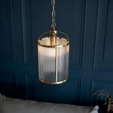 Endon Caldcombe pendant with cylindrical ribbed glass shade and antique brass frame, hanging from a chain against a dark blue panelled wall.