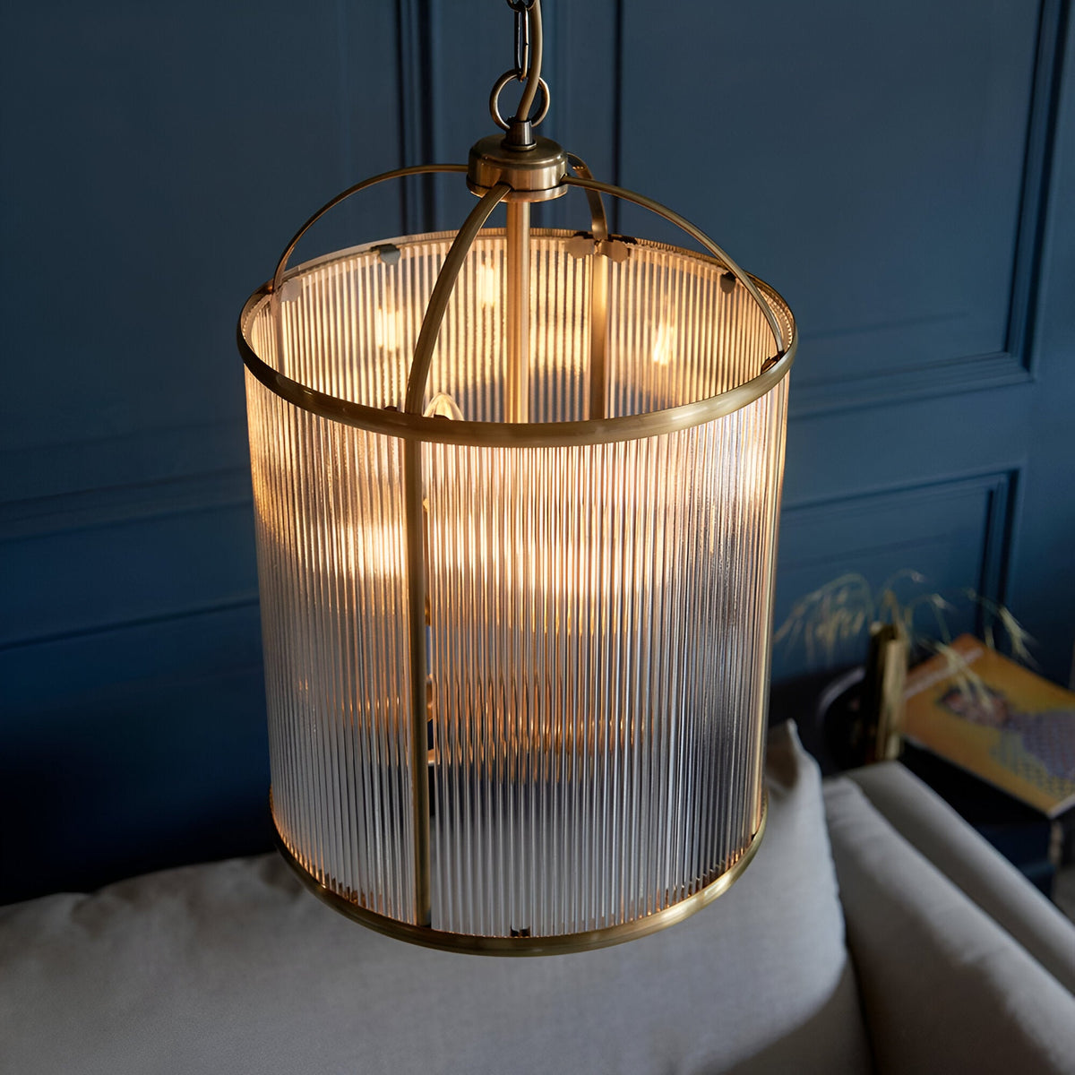Endon Caldcombe 4 light pendant with clear ribbed glass shade and antique brass frame, suspended from a chain against a dark blue panelled wall.