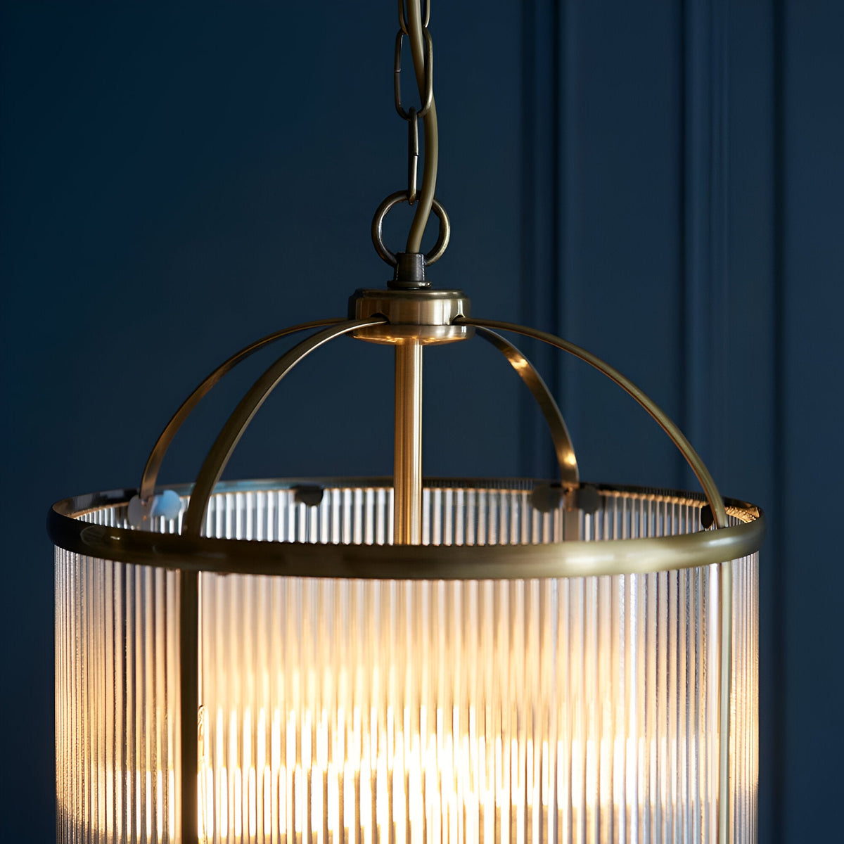 Close-up of Endon Caldcombe 4 light pendant showing antique brass domed frame, ribbed glass shade and chain suspension against a dark blue panelled wall.