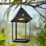 Brenmont Medium Table Lamp in matt black, bird-house lantern design with integrated LED, shown hanging outdoors from a tree branch amid greenery.