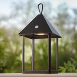 Brenmont Medium Table Lamp in matt black, a bird-house lantern with pyramid roof, open frame sides and top carry hoop, shown outdoors with warm LED glow.