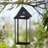 Endon Brenmont large matt black bird-house lantern table lamp hanging from a tree branch outdoors, illuminated with warm LED light amid green foliage.
