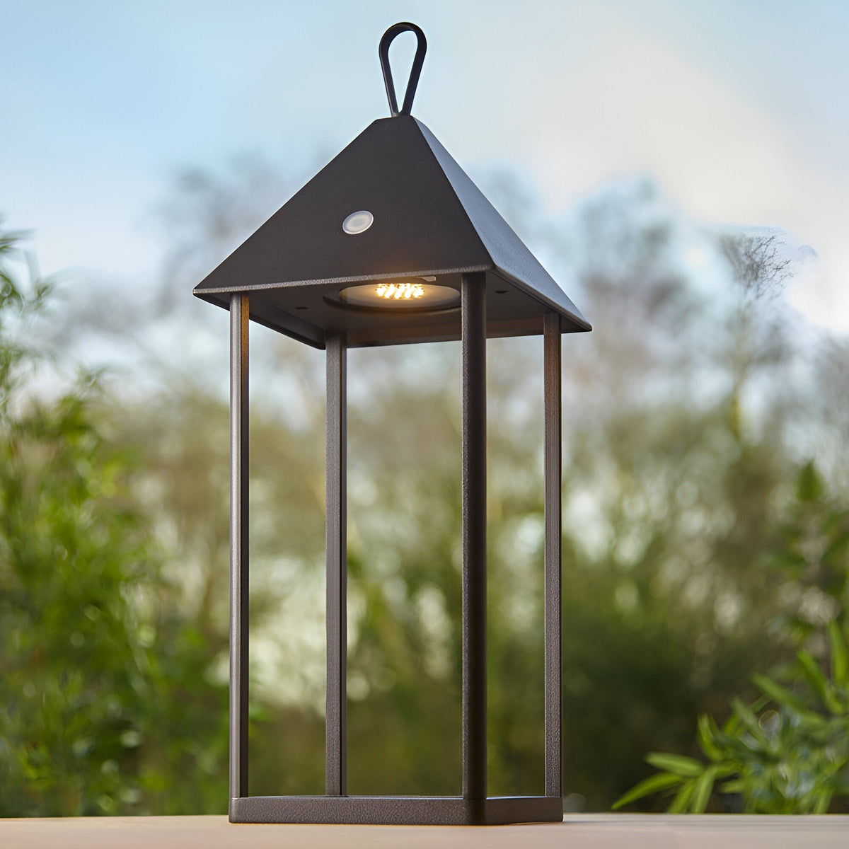 Endon Brenmont large table lamp in matt black, a modern bird-house lantern with pitched roof, loop handle and integrated LED, shown outdoors against foliage.