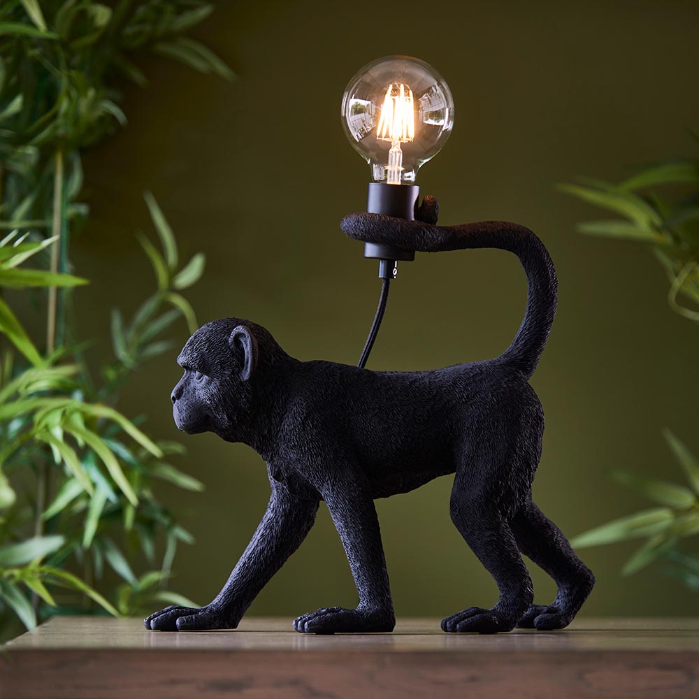 Belvoir Table Lamp in black resin, shaped as a walking monkey holding an exposed filament bulb, shown on a wood surface against greenery.
