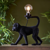 Belvoir Table Lamp in black resin, shaped as a walking monkey holding an exposed filament bulb, shown on a wood surface against greenery.