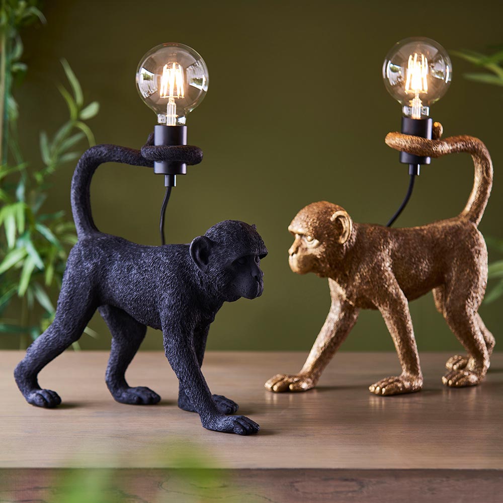 Belvoir Table Lamp in black, a resin monkey figure holding an exposed filament bulb, shown beside the gold version on a wooden surface.