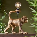Endon Belvoir table lamp shaped as a gold resin monkey holding an exposed filament bulb above its tail, shown on a shelf with foliage behind.
