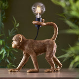 Endon Belvoir table lamp shaped as a gold resin monkey with tail curled around an exposed filament bulb holder, set on a wooden surface with foliage behind.