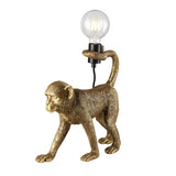 Endon Belvoir table lamp in gold resin, modelled as a walking monkey holding an exposed filament bulb in its tail, shown on a white background.