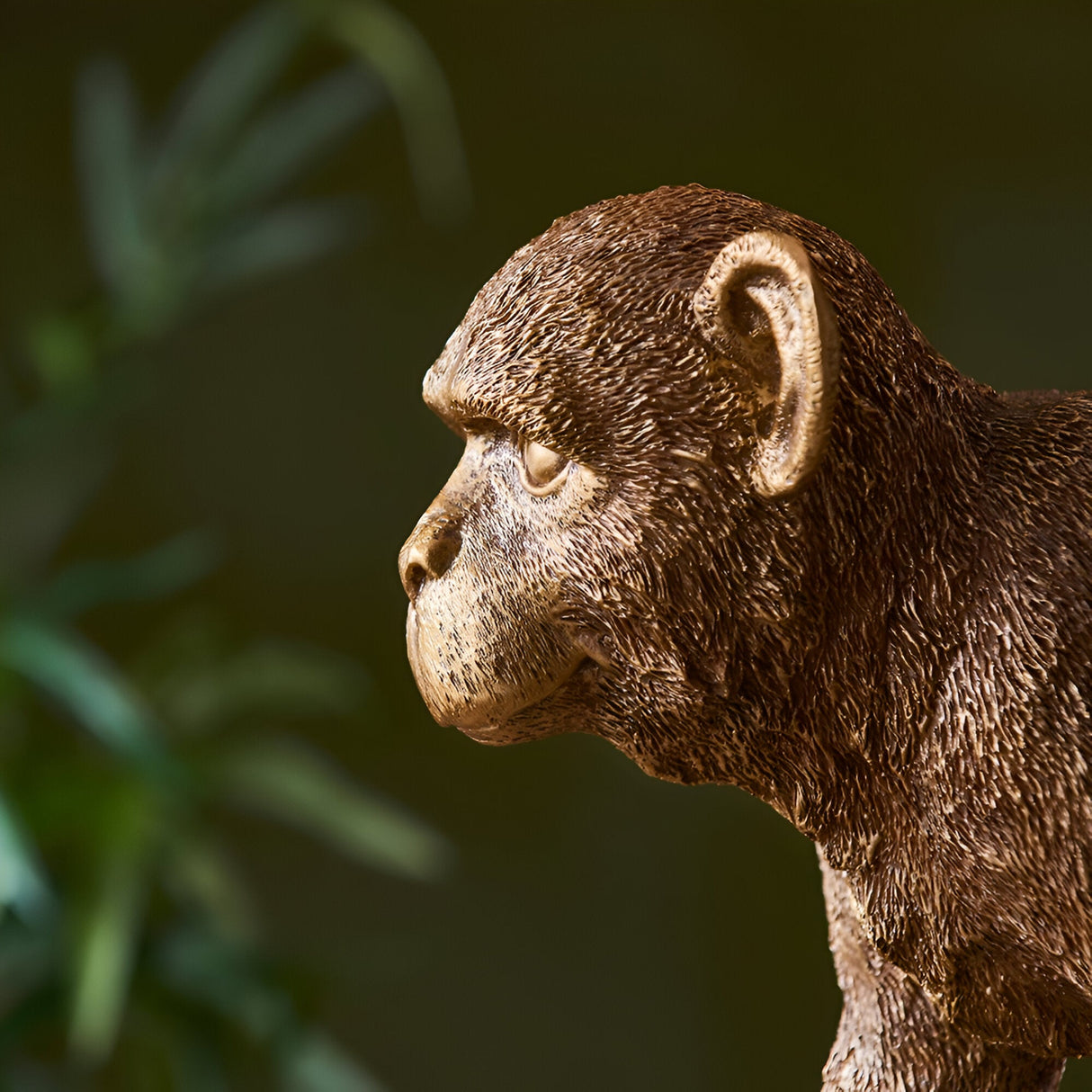 Endon Belvoir table lamp in gold, close-up profile of the detailed resin monkey figurine's head against a blurred green foliage background.