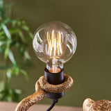 Close-up of the Endon Belvoir table lamp's exposed E27 filament globe bulb held in the gold resin monkey's hand against a green backdrop.