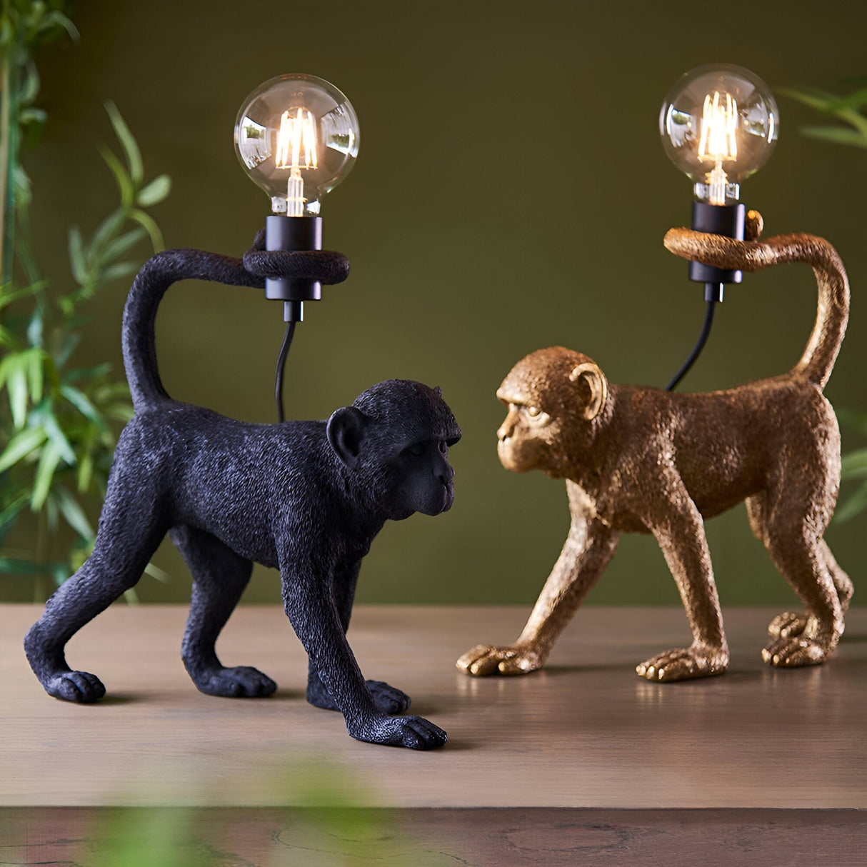 Endon Belvoir table lamps shown as matt black and vintage gold resin monkey figurines holding exposed filament bulbs on a wooden surface.