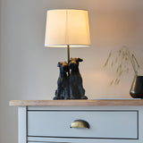 Danegrove Table Lamp in matt black resin, featuring two dog figures at the base and a cream fabric shade, shown on a wooden sideboard beside a dark vase.