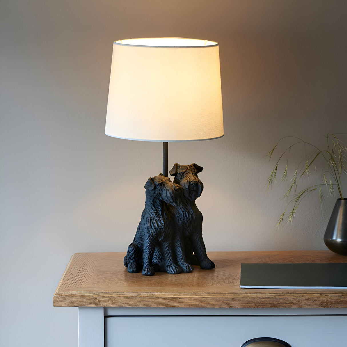 Danegrove Table Lamp in matt black resin, featuring two dogs at the base and a cream fabric shade, shown lit on a wooden sideboard.