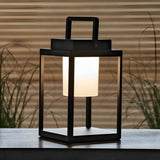 Endon Danedon portable table lamp in matt black with white polycarbonate diffuser and carry handle, lit on an outdoor table beside grasses.