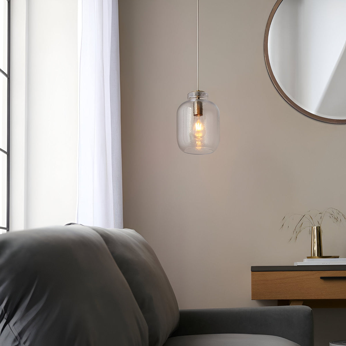 Caldwood Pendant with clear textured glass shade and antique brass fitting, hung above a grey armchair near a round mirror and wooden console.