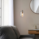 Caldwood Pendant with clear textured glass shade and antique brass fitting, hung above a grey armchair near a round mirror and wooden console.
