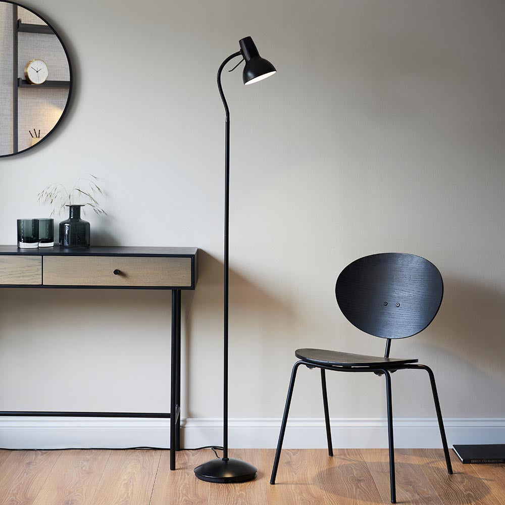 Endon Alderton matt black floor lamp with adjustable head and curved handle, beside a wooden console and black chair in a living room setting.