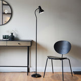 Endon Alderton matt black floor lamp with adjustable head and curved handle, beside a wooden console and black chair in a living room setting.