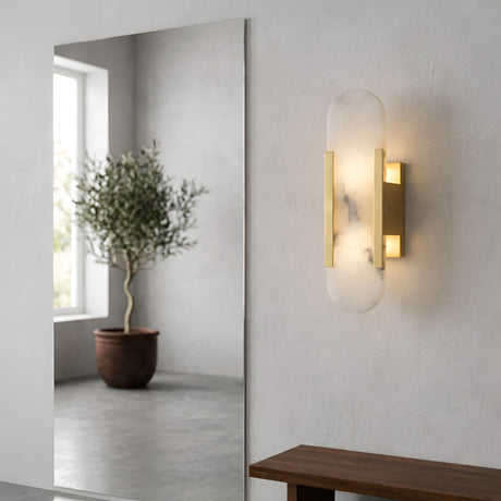 The Lirone 1 Light Small Vertical Capsule Alabaster Wall Light in soft white and brass is mounted on a light gray wall, with a large mirror reflecting a potted olive tree and a wooden bench placed in the foreground.