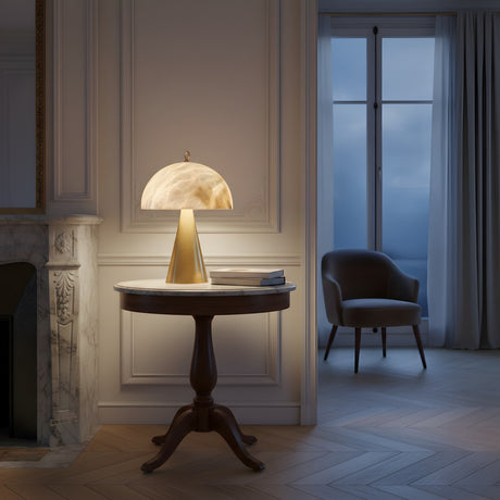 The Viren 2 Light Dome Crown Alabaster Table Lamp in brushed bronze and soft white sits on a round wooden table near a window with drawn curtains, casting ambient light in an elegant room with a fireplace and an armchair.