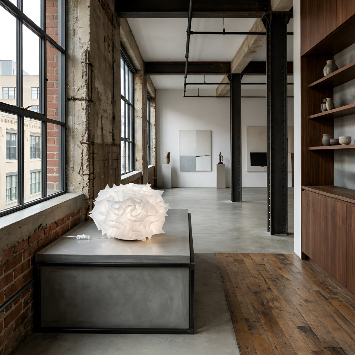 A modern art gallery with concrete floors, brick walls, large windows, and the Violla 1 Light Table Light - White on a gray pedestal. Minimalist paintings, mood lighting, and shelves with stylish decor complete the scene.