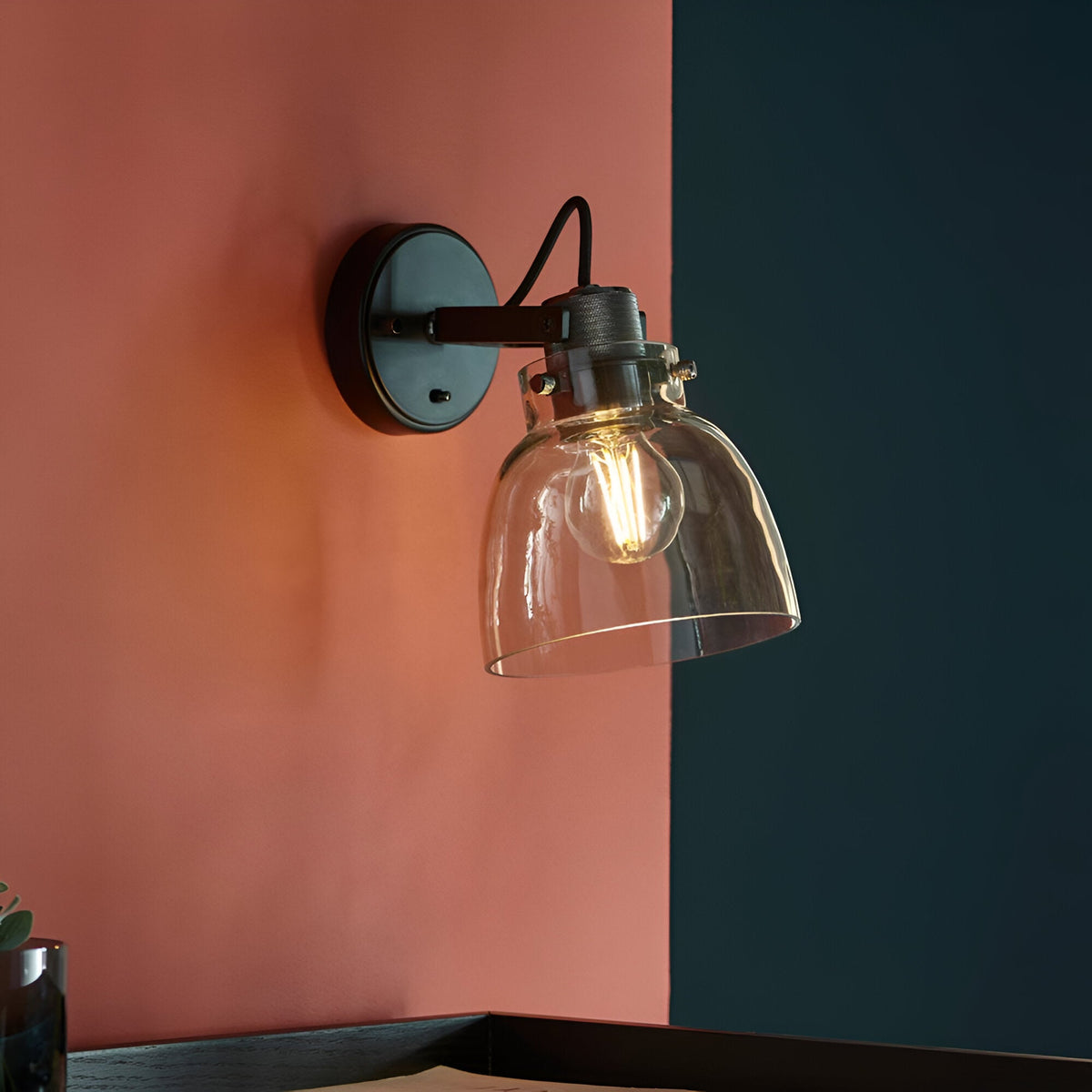 Camden Wall Light in black and chrome with clear glass dome shade and exposed bulb, mounted on a coral wall beside a dark blue accent wall.