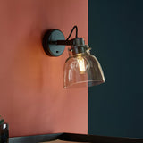 Camden Wall Light in black and chrome with clear glass dome shade and exposed bulb, mounted on a coral wall beside a dark blue accent wall.