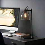 Endon Camden industrial task table lamp in matt black with knurled details and smoked glass shade, on a desk beside a monitor, keyboard and glasses.
