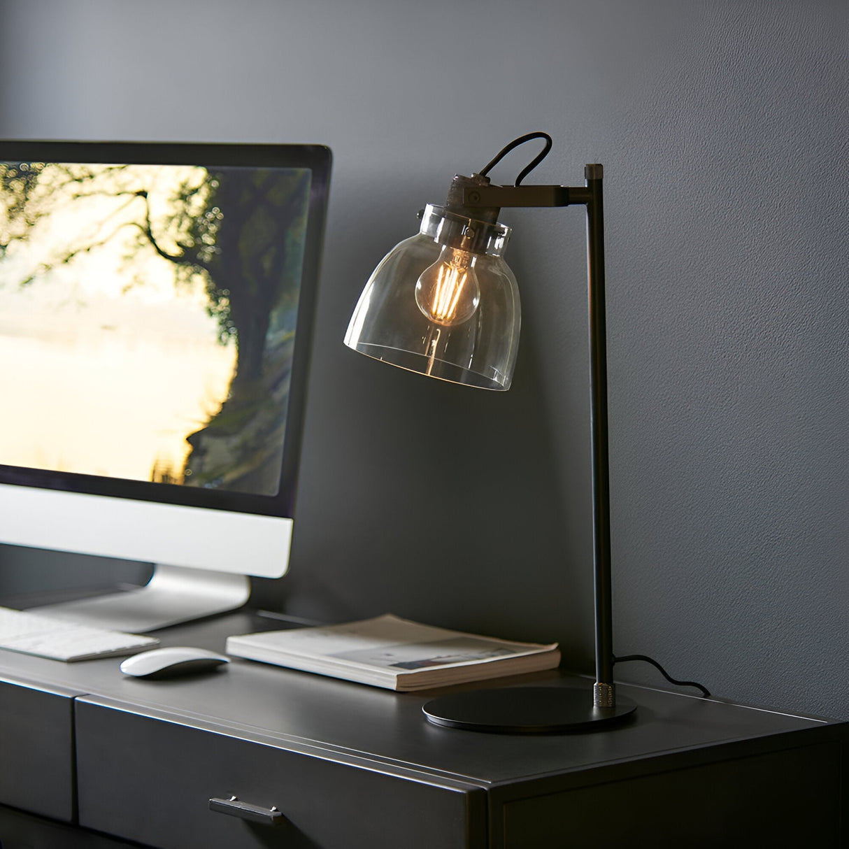 Endon Camden industrial task table lamp in matt black with knurled details and smoked glass shade, lit on a desk beside a monitor and keyboard.