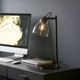 Endon Camden industrial task table lamp in matt black with knurled details and smoked glass shade, lit on a desk beside a monitor and keyboard.